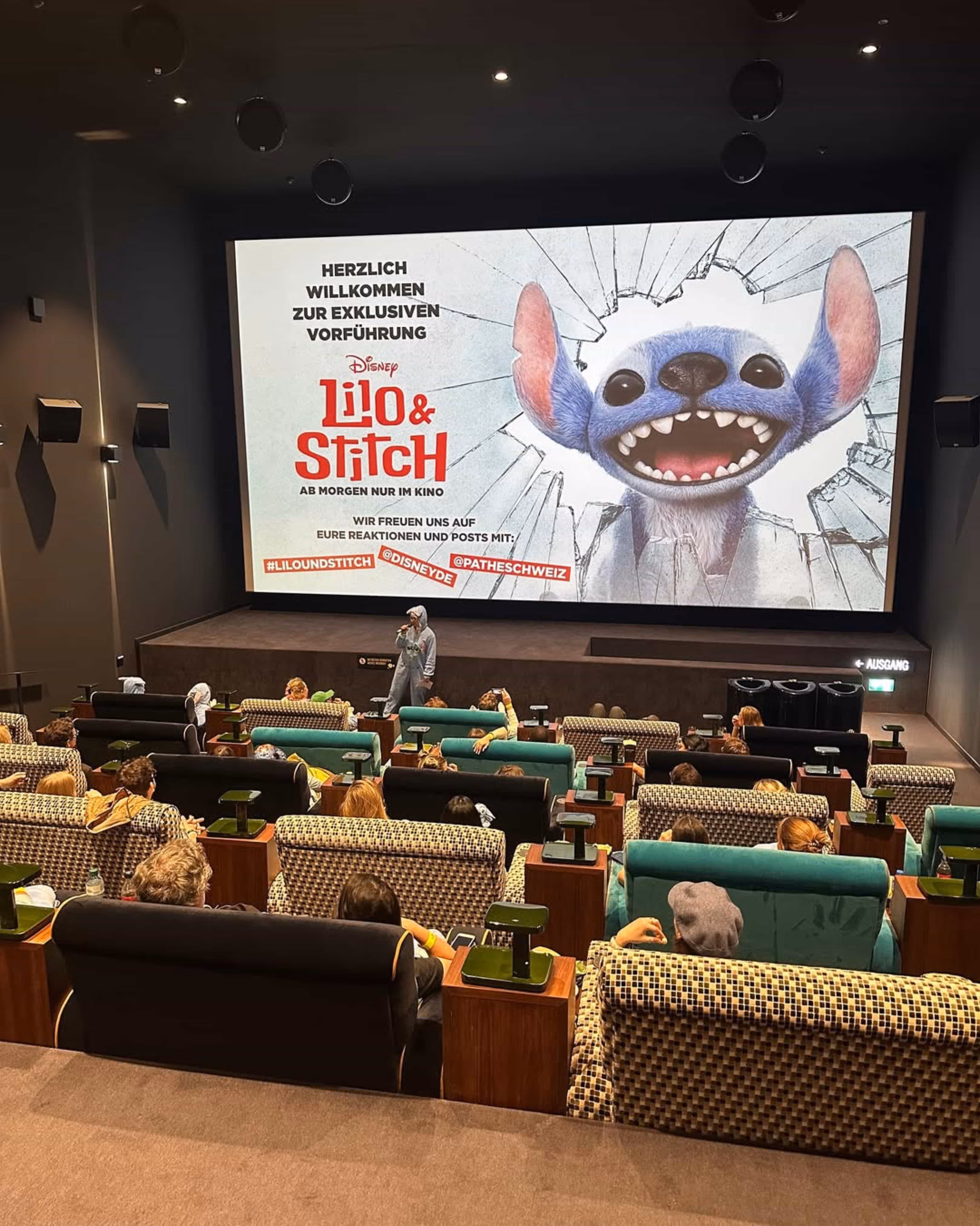 Person in Stitch-Kostüm spricht vor Publikum in einem Kinosaal mit Lilo & Stitch Vorführungsbild auf der Leinwand.