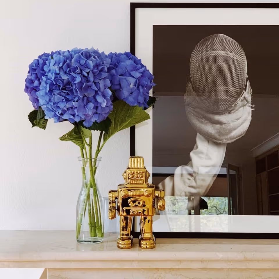 Eine Vase mit blauen Hortensien neben einer goldfarbenen Roboterfigur vor einem gerahmten Foto einer Fechtausrüstung.