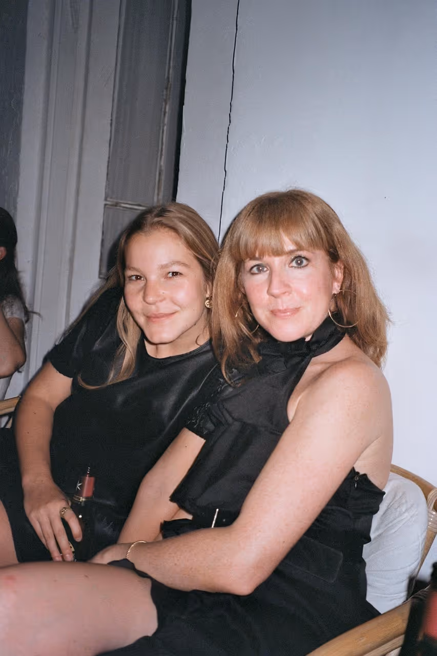 Zwei Frauen in schwarzer Kleidung sitzen eng nebeneinander auf Stühlen und lächeln in die Kamera.