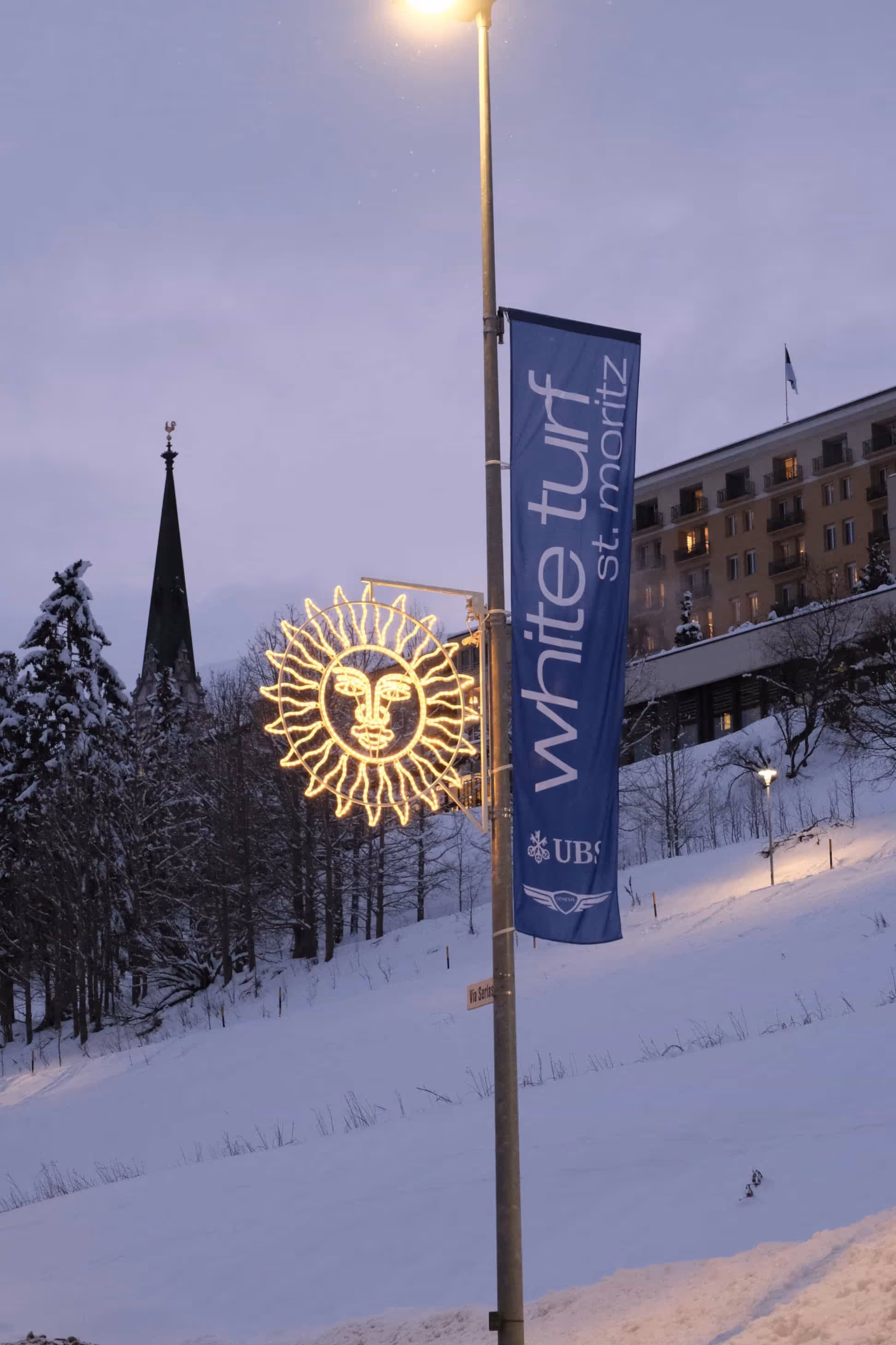 Beleuchtete Sonnenskulptur und Flagge mit Aufschrift 'white turf st. moritz' an einem Laternenpfahl in verschneiter Landschaft bei Dämmerung.