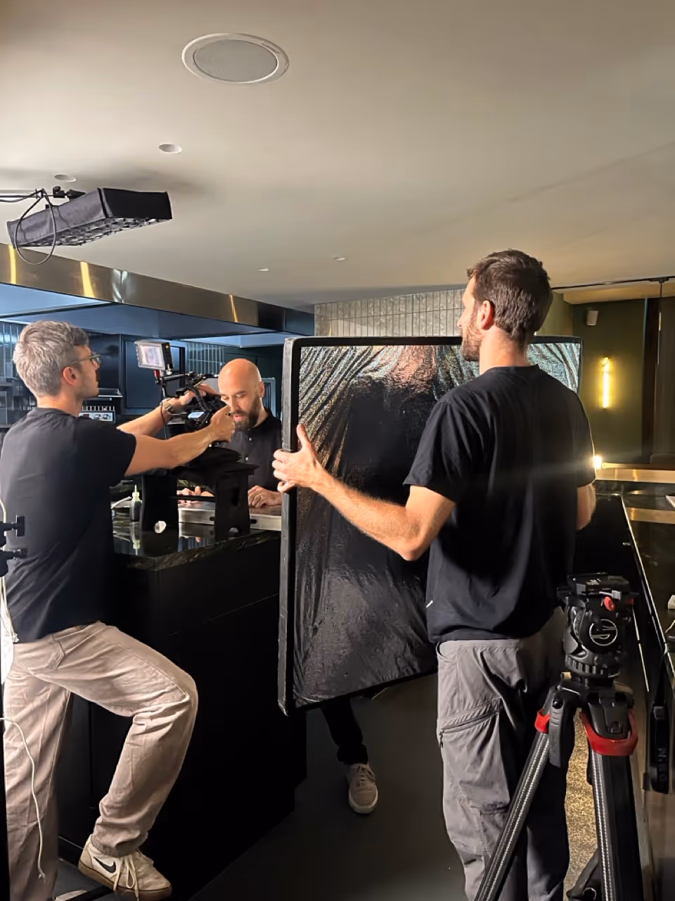 Zwei Männer bedienen Kamera- und Lichtausrüstung, während ein dritter Mann an einem schwarzen Tresen steht und nach unten schaut.