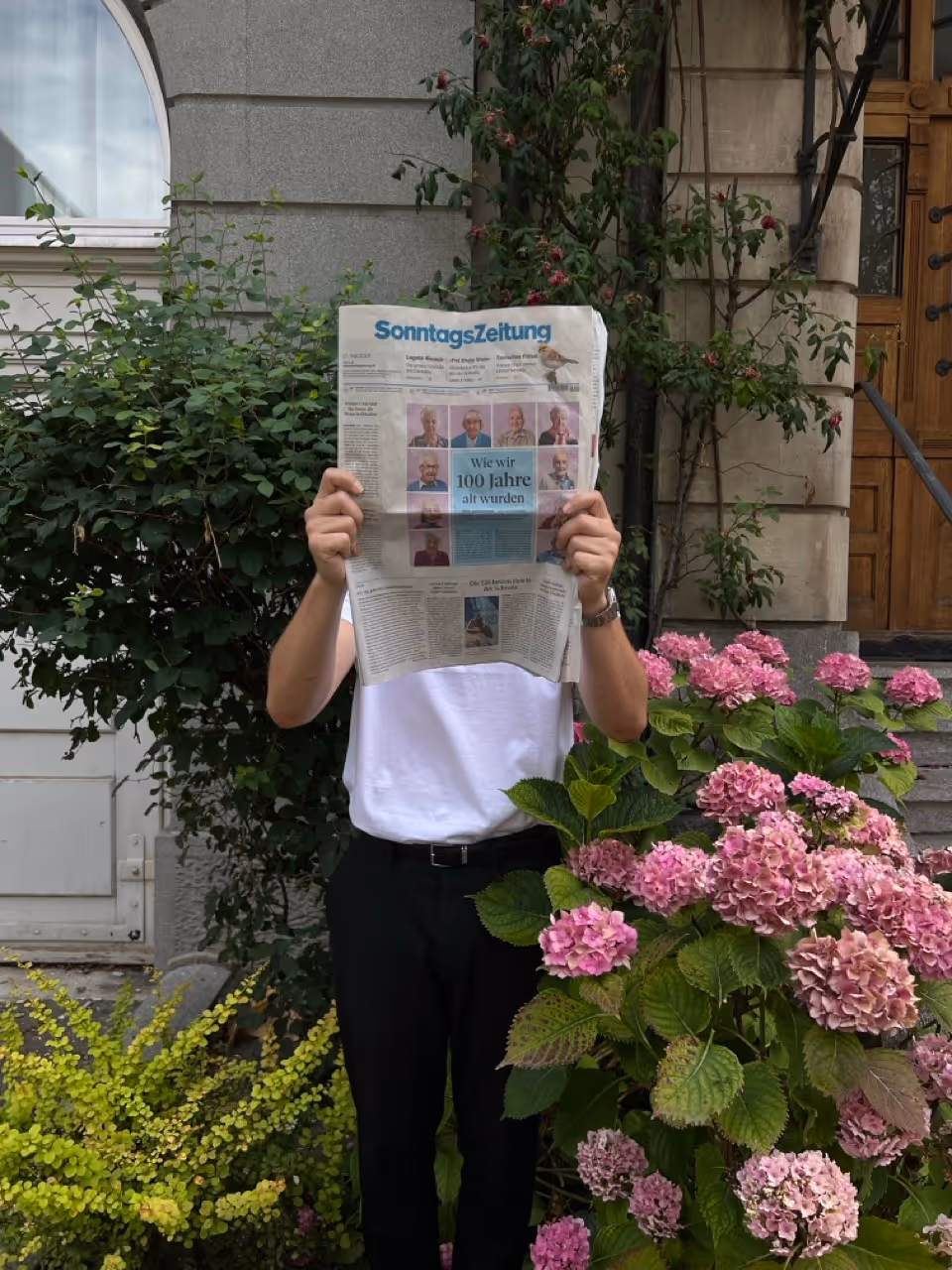 Person trägt weisses T-Shirt und schwarze Hose, hält vor dem Gesicht eine Ausgabe der SonntagsZeitung in einem Garten mit pinkfarbenen Blumen.