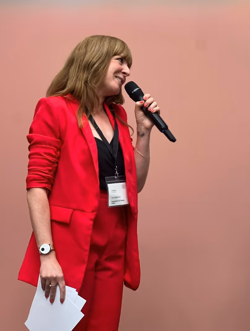 Frau in rotem Anzug hält Mikrofon und Papiere, spricht vor rosa Hintergrund.