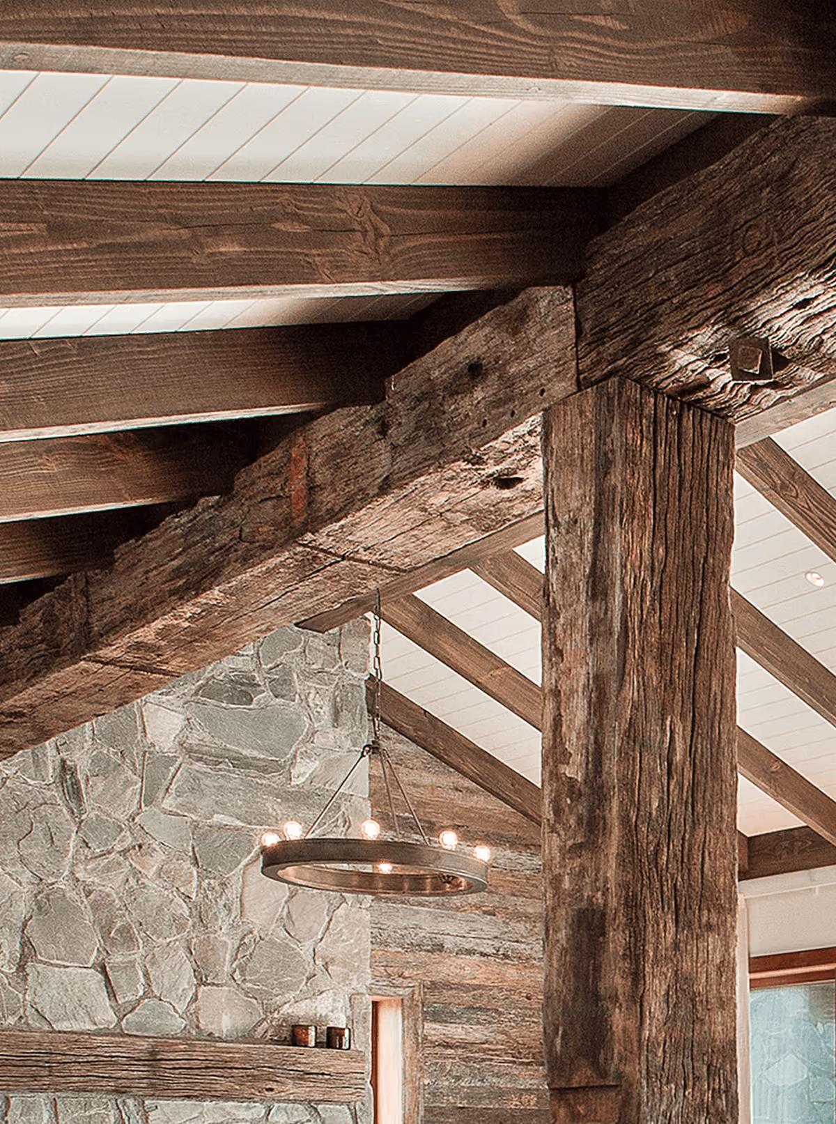 Interior of a rustic room featuring exposed dark wooden beams and posts supporting a white-paneled ceiling, with a stone wall and a round chandelier with small lights hanging from the ceiling.