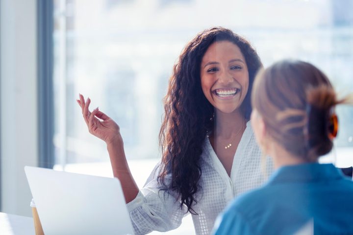 Smiling woman with curly hair talking to another person in a bright office setting with a laptop in front of her.
