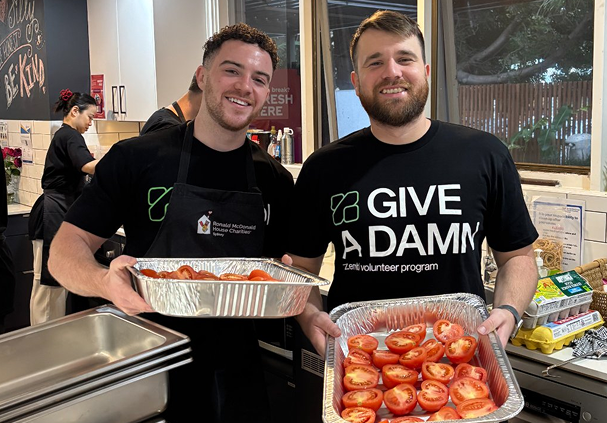 Two men smiling in a kitchen holding trays filled with halved tomatoes, one wearing a GIVE A DAMN t-shirt and the other a Ronald McDonald House Charities apron.