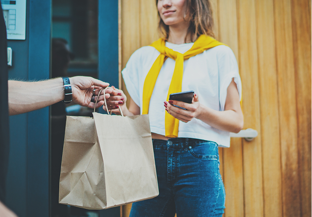 Person receiving two brown paper bags from another person while holding a smartphone near a wooden door.
