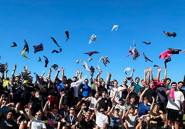 A large group of people outdoors throwing scarves or bandanas into the air against a clear blue sky.
