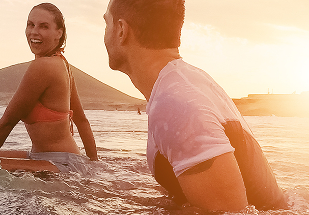 Man and woman sitting in shallow ocean water at sunset, with a hill in the background.