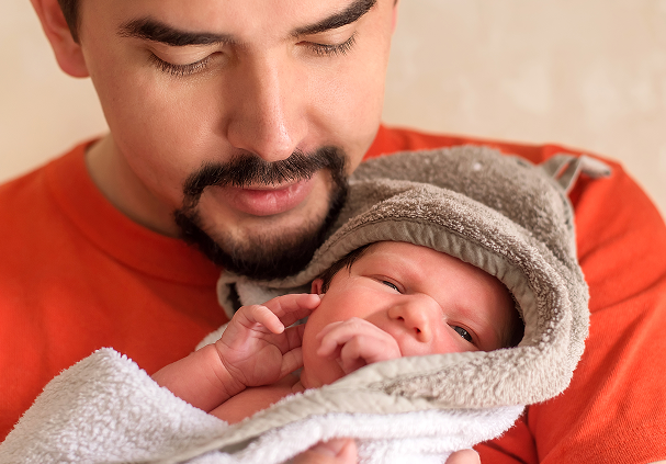 A man in an orange shirt tenderly holding a newborn baby wrapped in a towel with a hood.