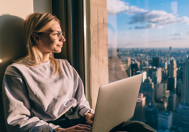 Woman wearing glasses sitting by a window with a laptop, looking out over a cityscape at sunset.