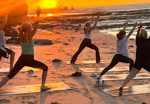 Group of people practicing yoga on mats on a sandy beach at sunset with the ocean in the background.