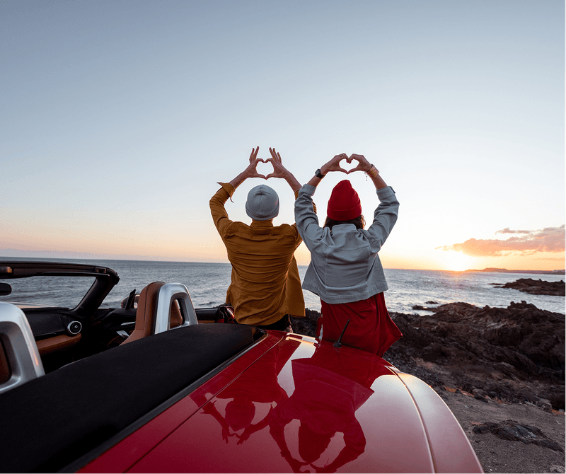 Two people sitting by the shore at sunset, making heart shapes with their hands above their heads next to a red convertible car.
