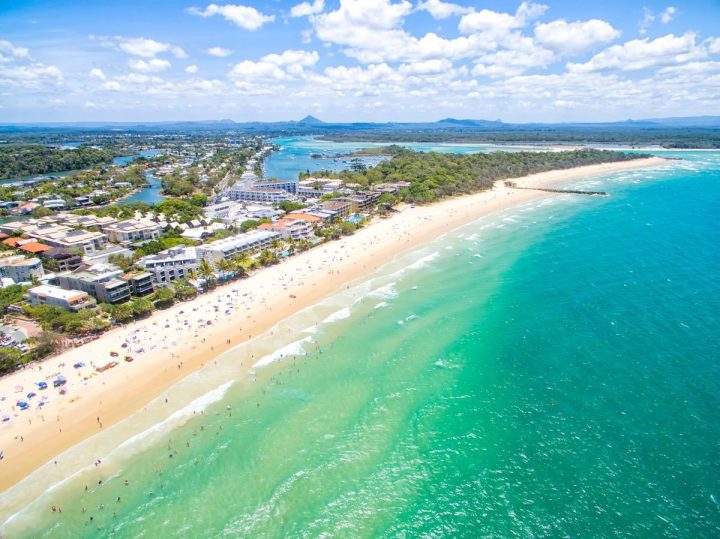 Aerial view of a sunny beach with turquoise water, sandy shore, and buildings along the coastline under a partly cloudy sky.