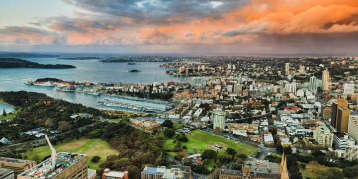 Aerial view of a coastal city at sunset featuring a large harbor with boats and densely packed buildings surrounded by green parks.