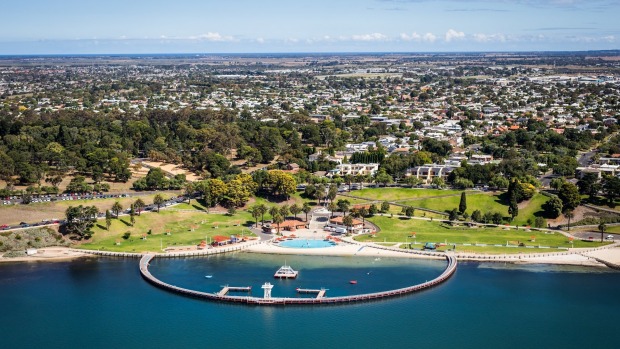 Aerial view of a coastal park with a curved pier enclosing a swimming area and a city in the background.