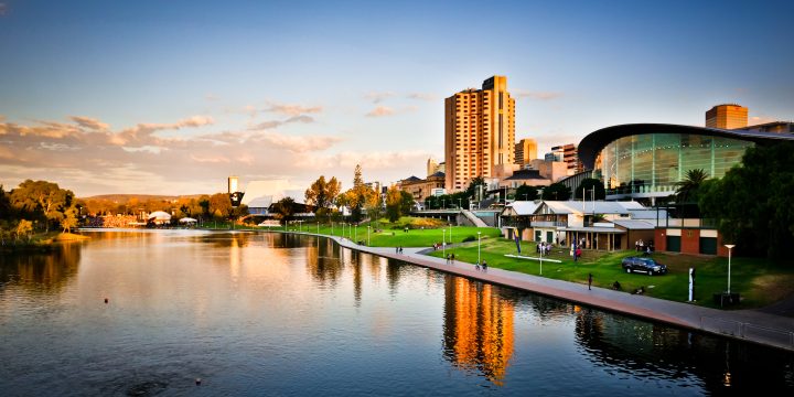 City skyline at sunset with modern buildings reflecting on a calm river and a riverside walkway with greenery.