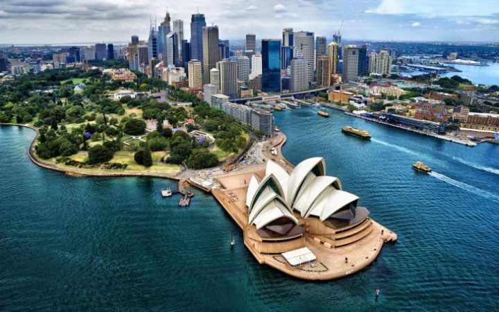 Aerial view of Sydney Opera House on the harbor with city skyline and green parkland in the background.