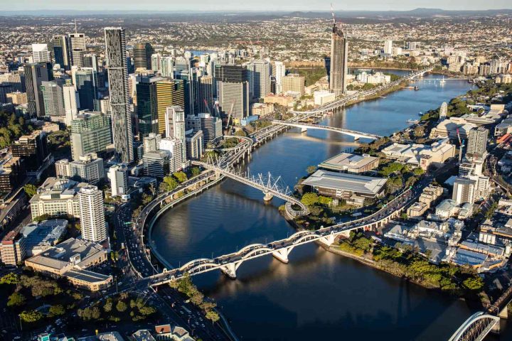 Aerial view of a city skyline with several bridges crossing a wide river, surrounded by tall buildings and urban areas under a clear sky.