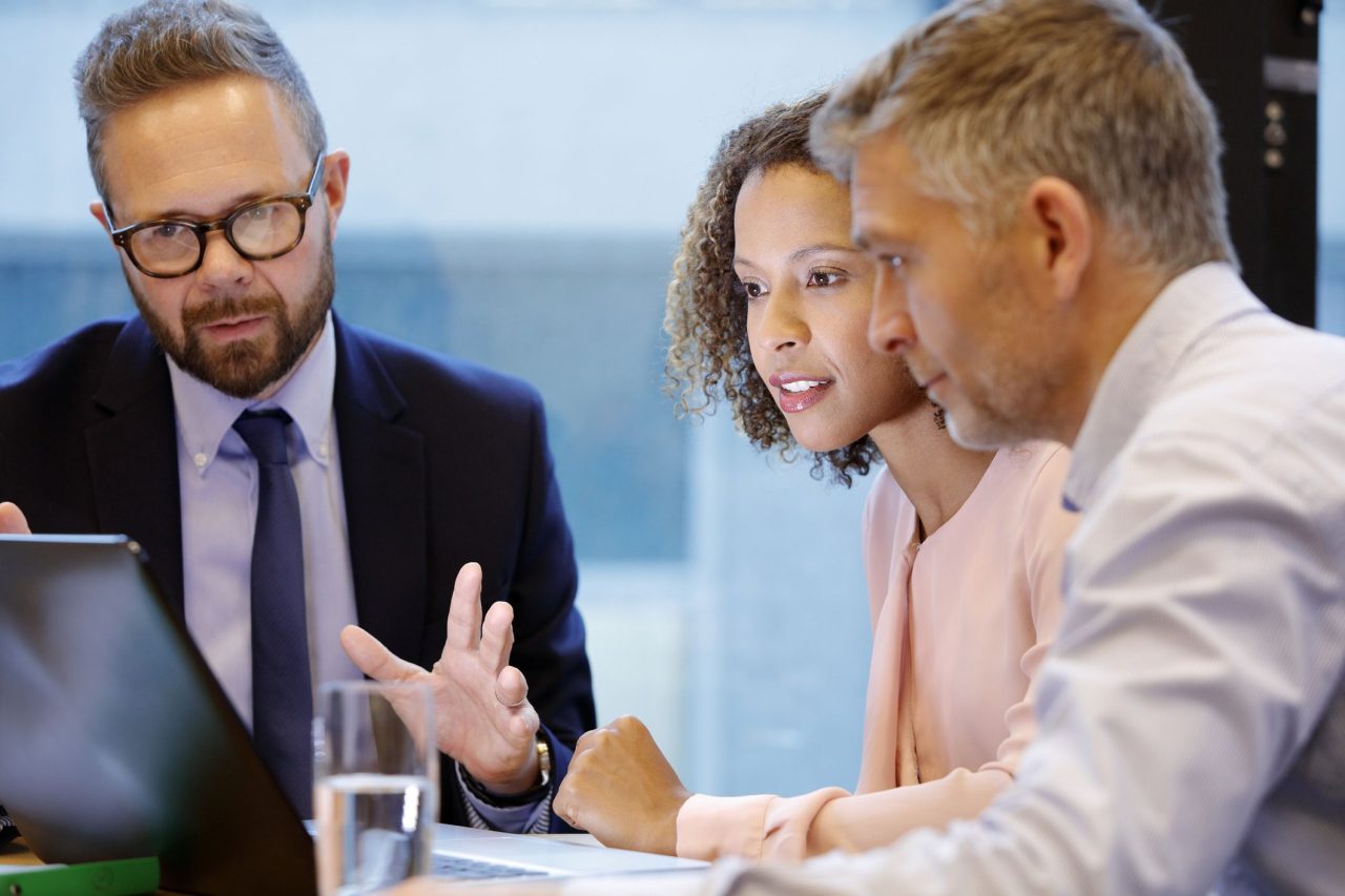 Three business professionals having a discussion while looking at a laptop screen in an office setting.