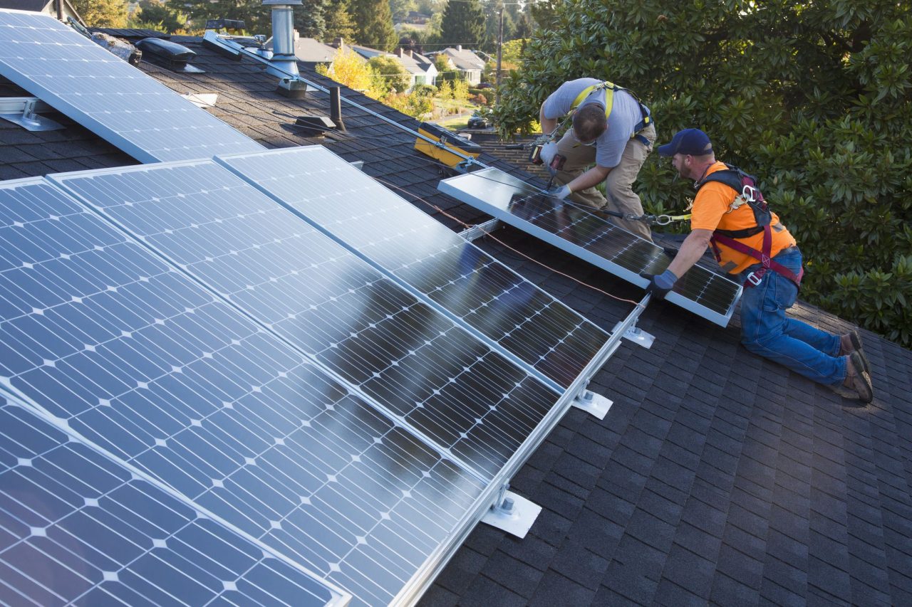 Two workers wearing safety harnesses installing solar panels on a residential roof.