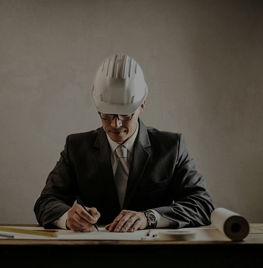 Man wearing a white hard hat and suit writing on a desk with architectural tools and blueprints.
