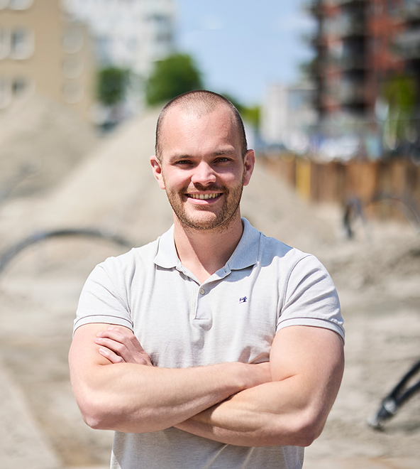 Smiling bald man with crossed arms wearing a light gray polo shirt, standing outdoors in front of a blurry construction site.