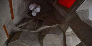 Interior view of a modern architectural space with angular concrete walls and ceiling featuring an abstract geometric light fixture and a red skylight.