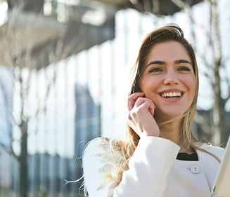 Smiling young woman in a white coat talking on a smartphone outdoors with a modern building in the background.