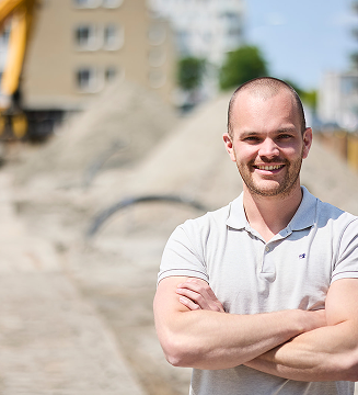 Smiling man with arms crossed standing outdoors in front of blurred construction site.