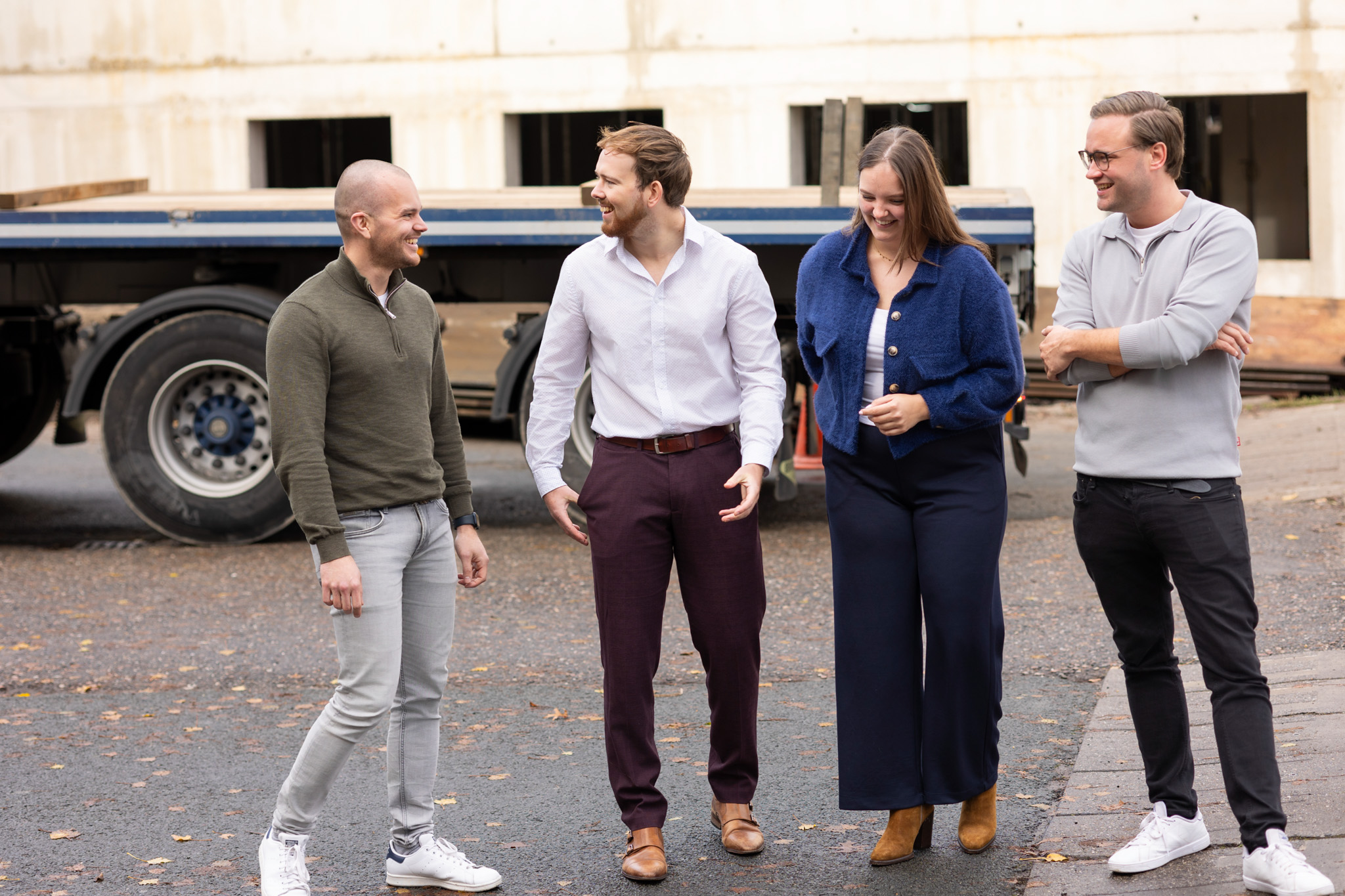 Four casually dressed people standing outdoors on a paved area near a truck, smiling and talking together.