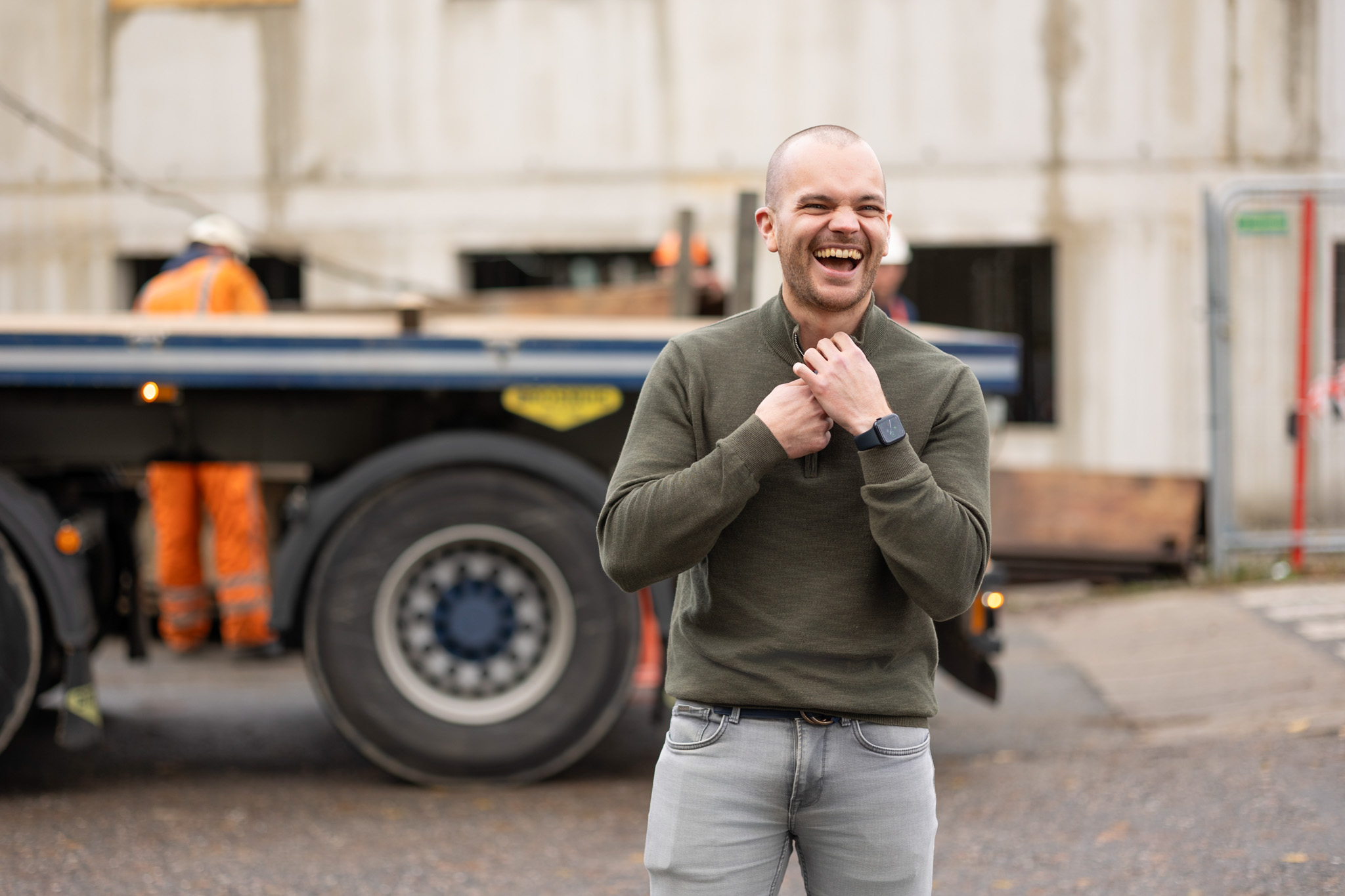 Smiling man in green sweater and gray jeans standing outdoors in front of a truck and construction workers.