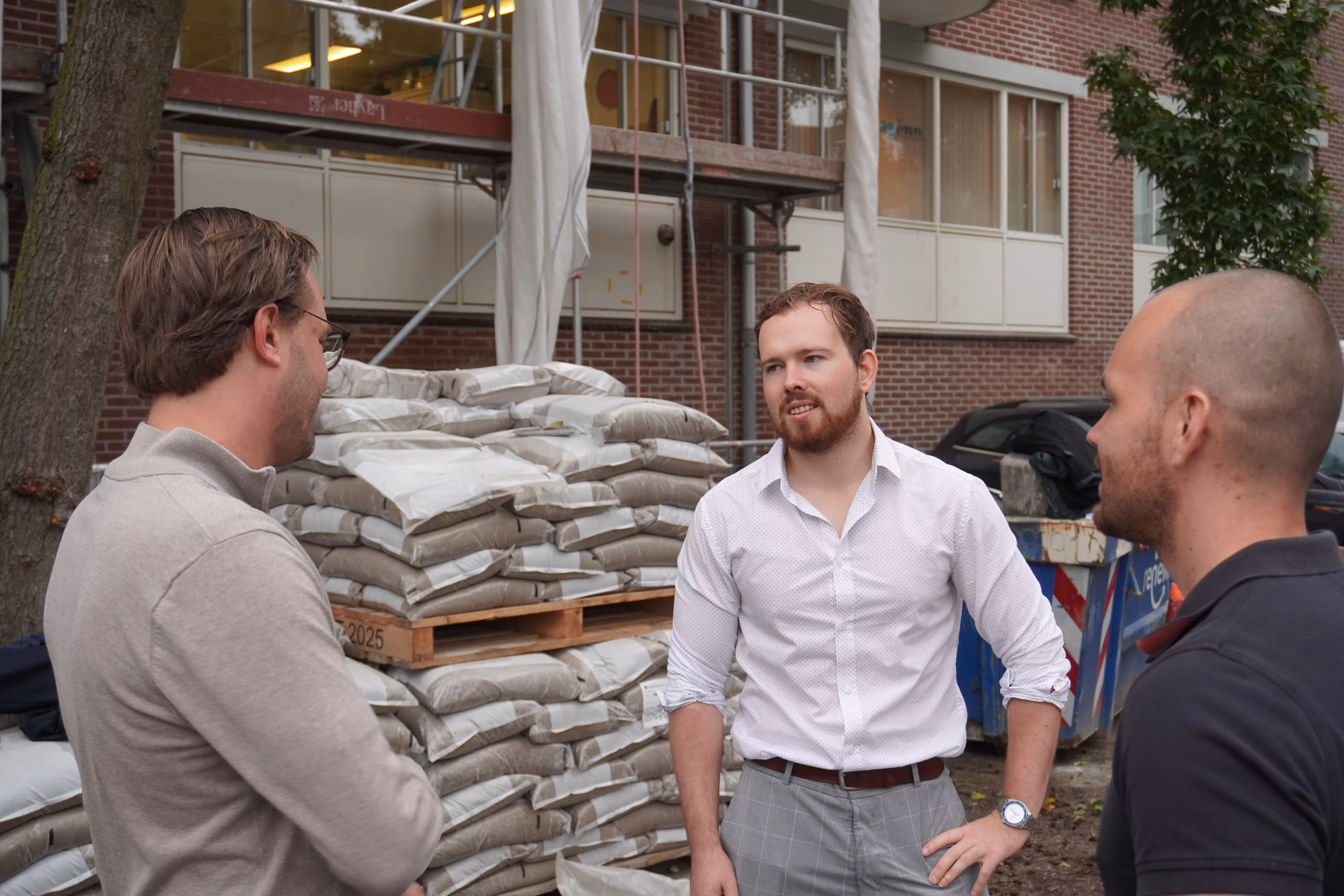 Three men standing outdoors in front of stacked bags on pallets, engaged in conversation near a brick building.