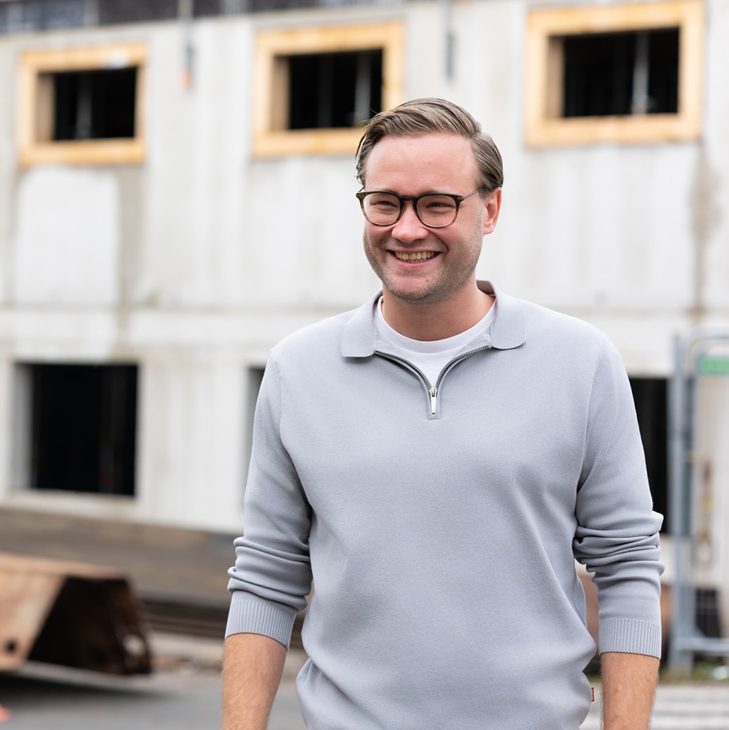 Smiling man with glasses wearing a light gray zip-up sweater standing outdoors in front of a building under construction.