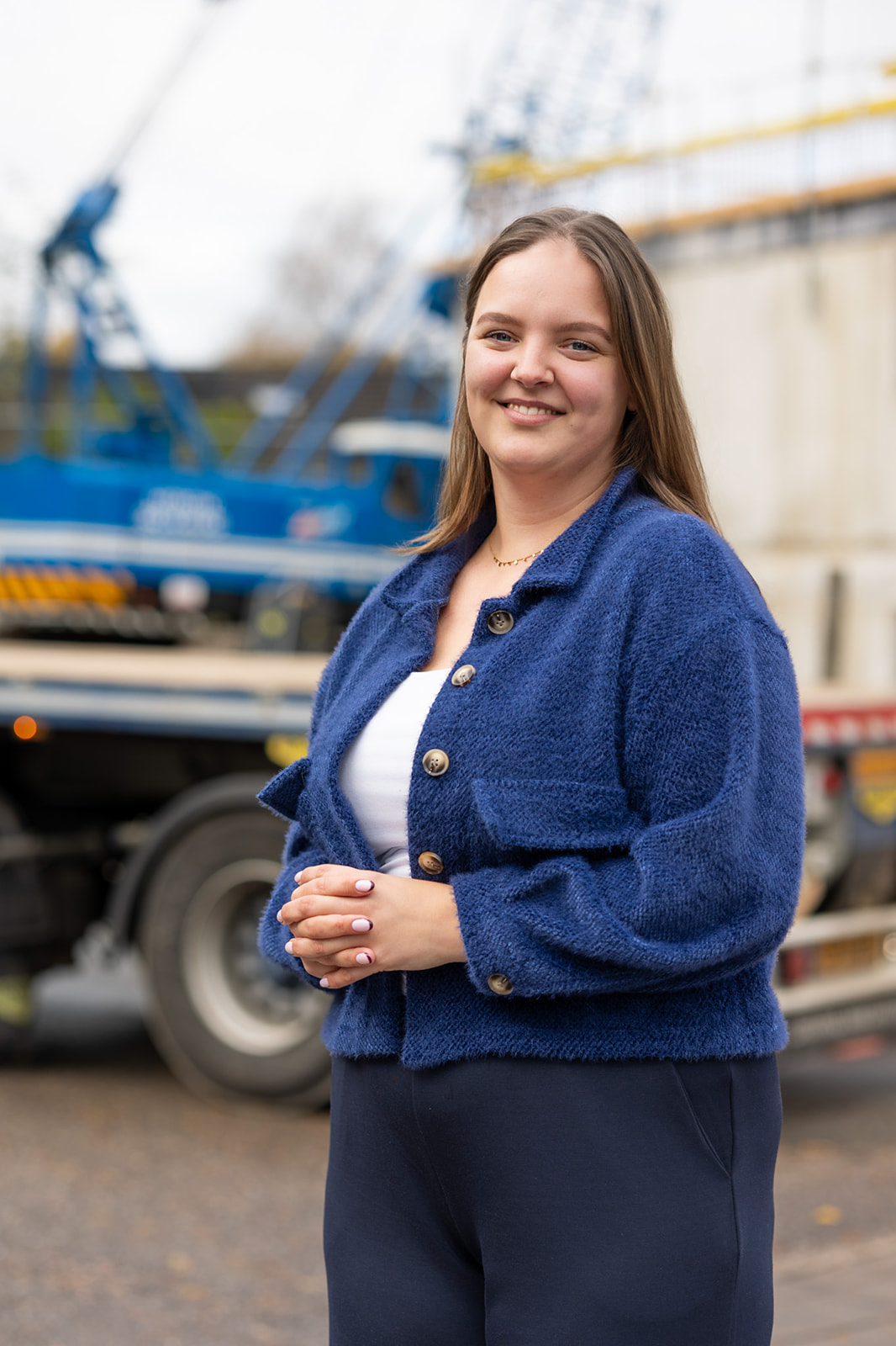 Smiling woman wearing a blue cardigan and white top standing outdoors with construction equipment in the background.