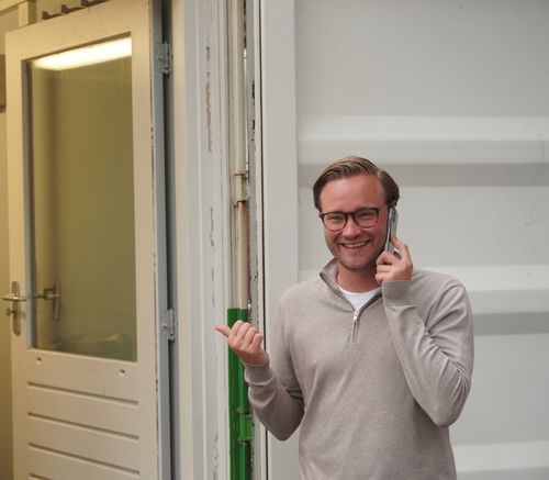 Smiling man wearing glasses and a beige sweater talking on a phone and pointing to an open door.