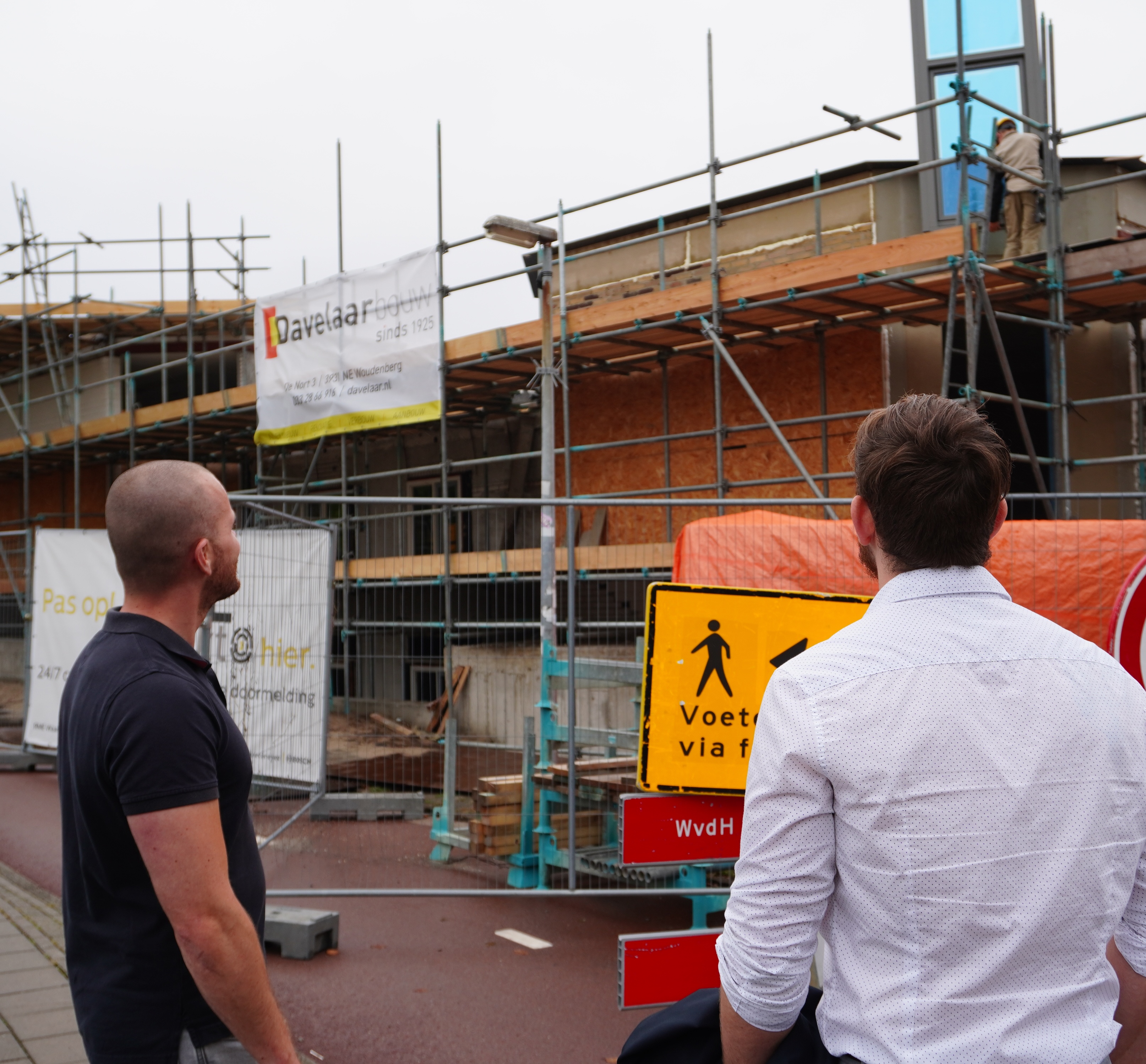 Two men, one in a black polo and one in a white shirt, stand on a street looking at a building under construction with scaffolding.