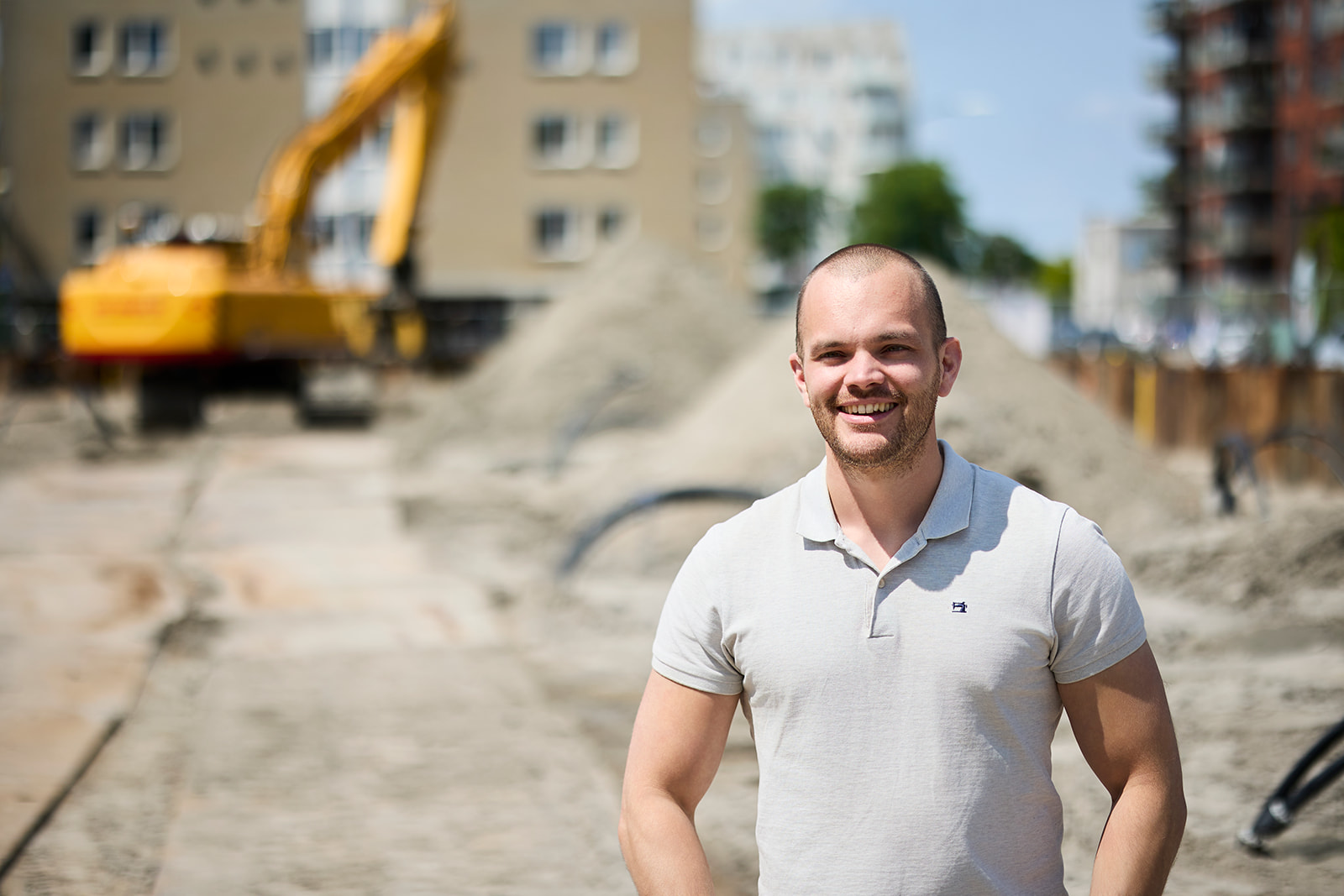 Smiling man in a white polo shirt standing at a construction site with an excavator and piles of dirt in the background.
