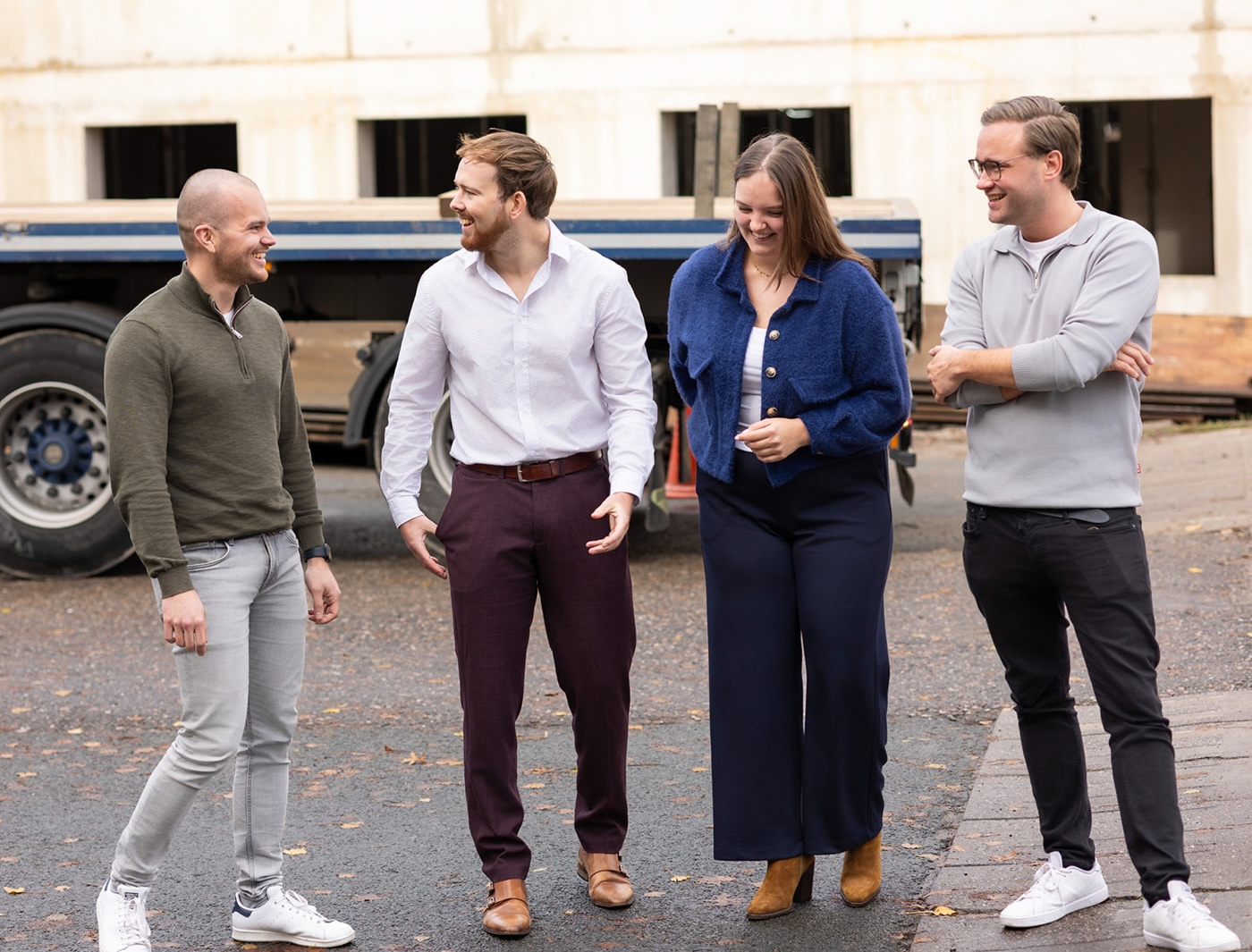 Four casually dressed people standing outdoors on pavement, smiling and talking near a large vehicle.