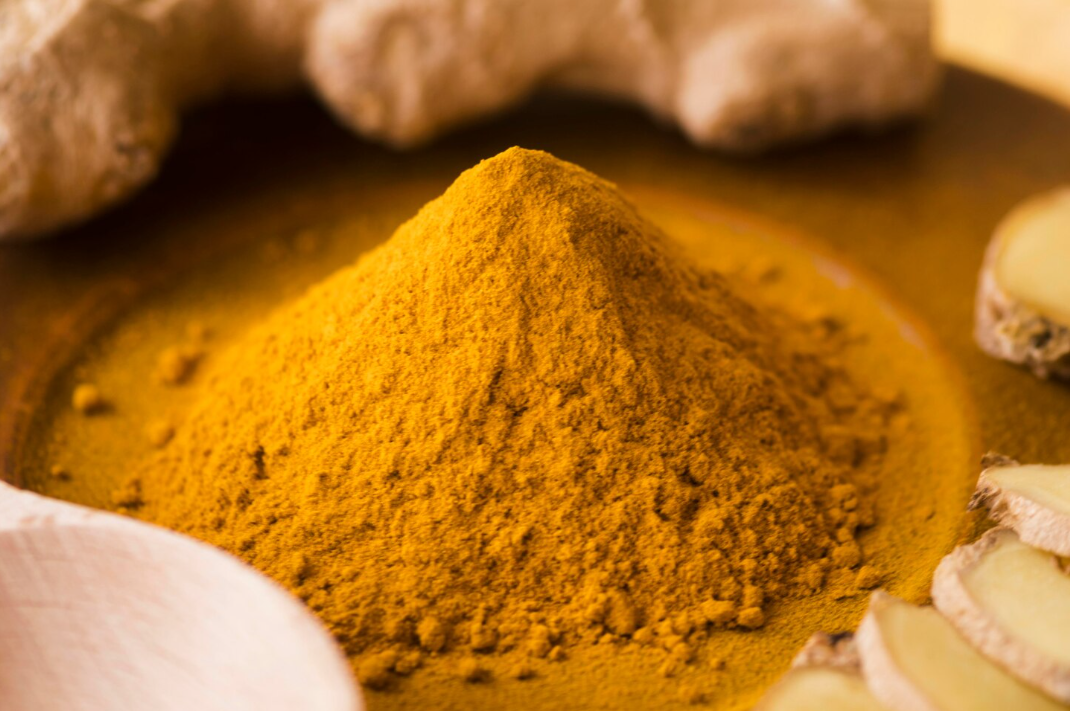 Pile of bright yellow turmeric powder on a wooden surface with fresh turmeric roots and slices nearby.