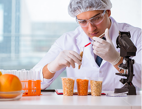 Scientist in lab coat and safety gear examining samples in test tubes next to microscope on lab table.