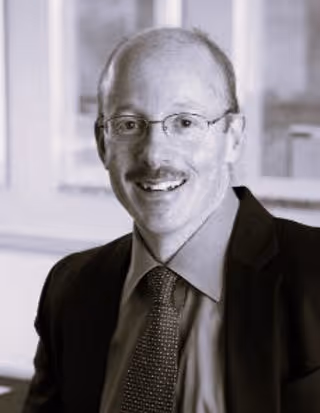 Smiling man with glasses, mustache, suit, and patterned tie in an office setting.