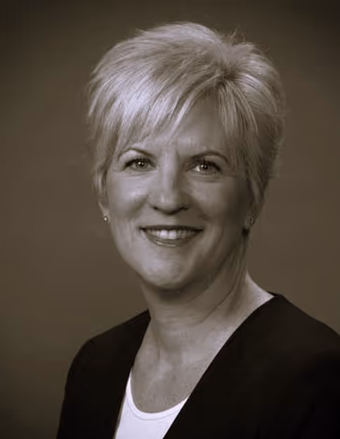 Smiling woman with short light hair wearing a dark blazer and white top against plain background.