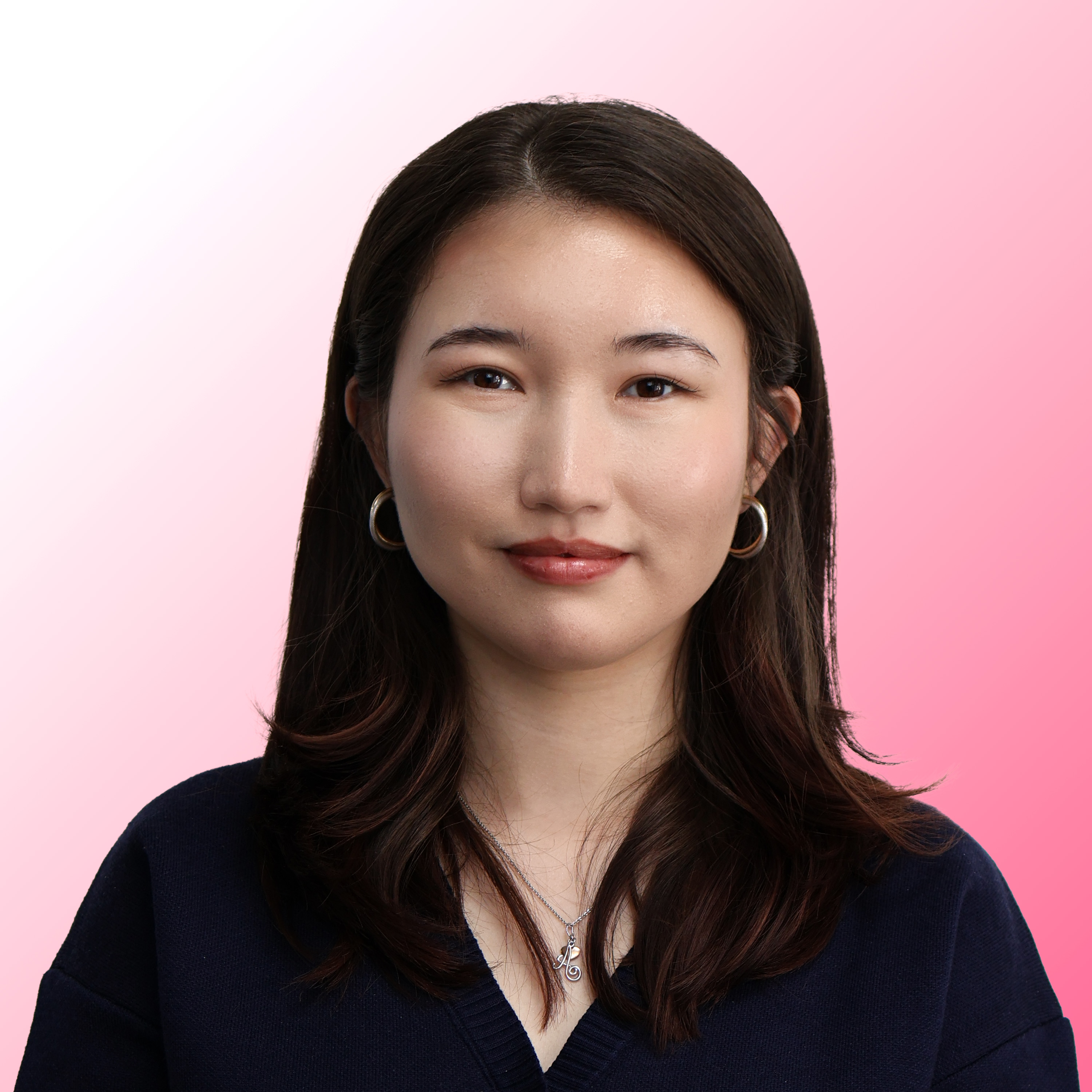 Portrait of a young woman with straight dark brown hair wearing hoop earrings, a silver necklace with a letter A pendant, and a dark navy V-neck sweater against a soft pink gradient background.