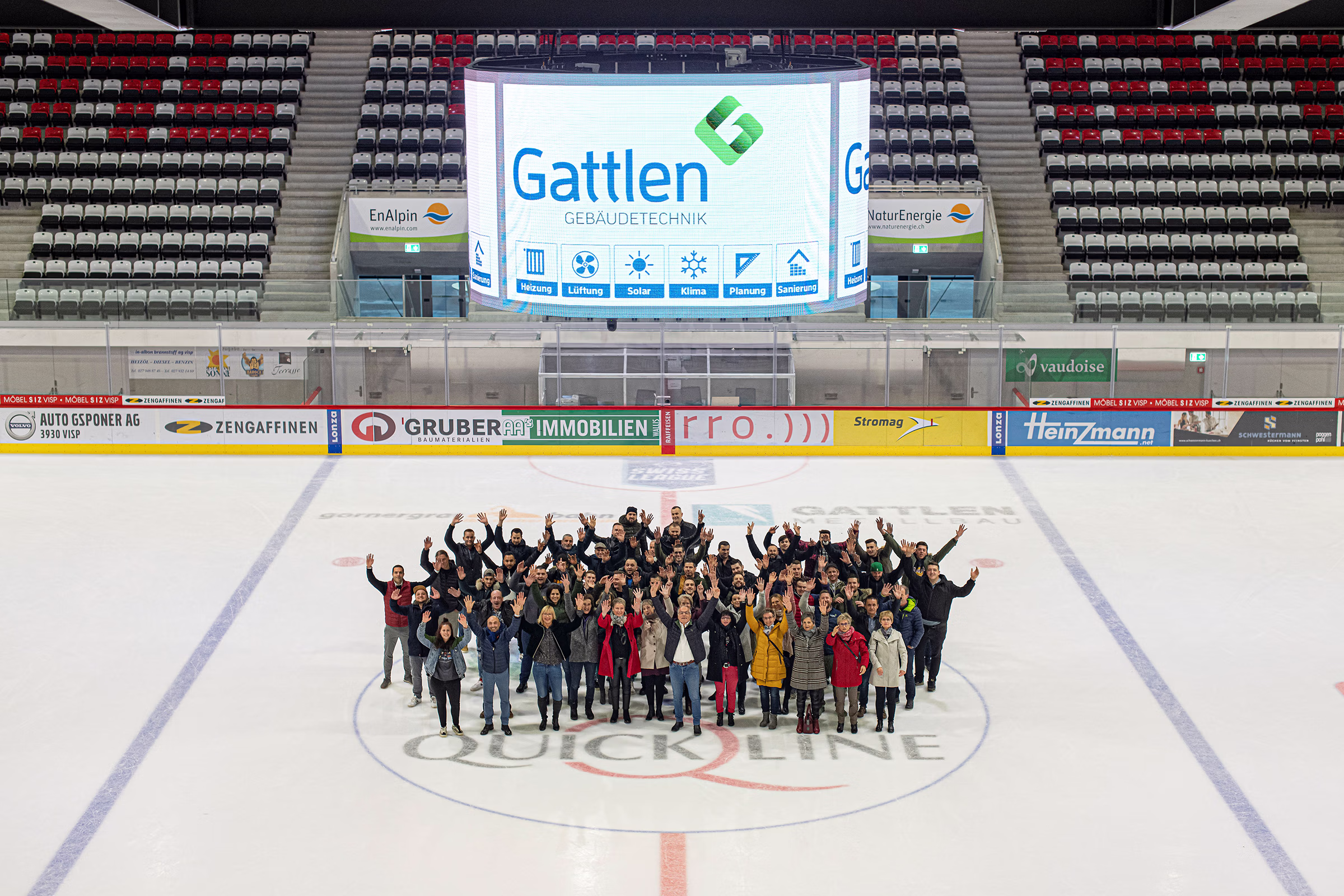 Gruppenfoto des gesamten Gattlen-Teams in der Lonza Arena Visp – rund 50 Mitarbeiter posieren gemeinsam auf dem Eis der Eishalle, im Hintergrund Sponsorenbanner und Firmenlogo