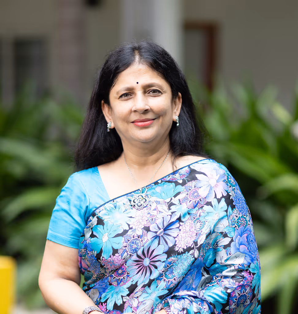 Middle-aged woman with black hair wearing a blue floral saree smiling in an outdoor setting.