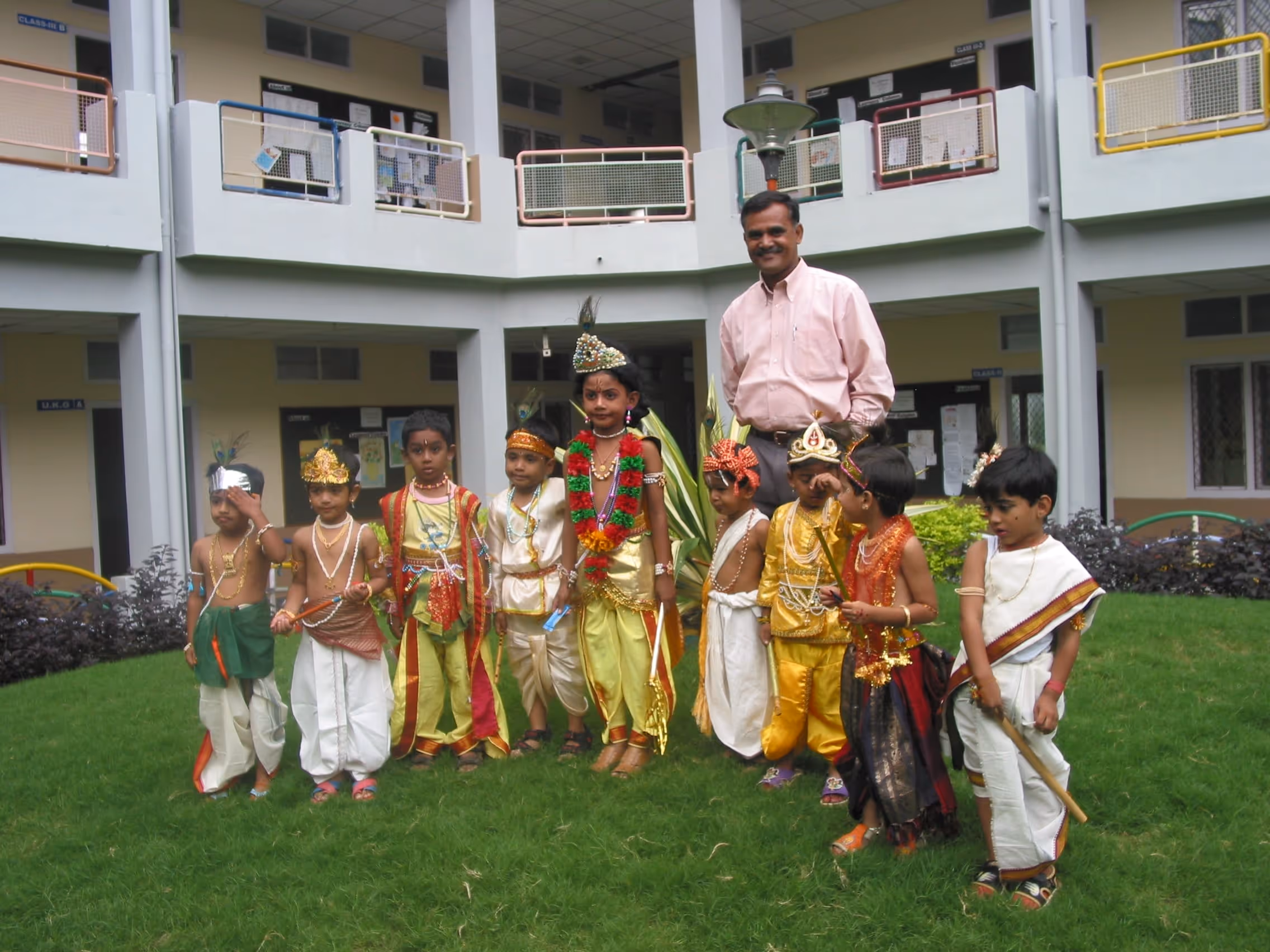 Group of young children dressed in traditional Indian costumes standing on grass with a man in a pink shirt behind them in a school courtyard.