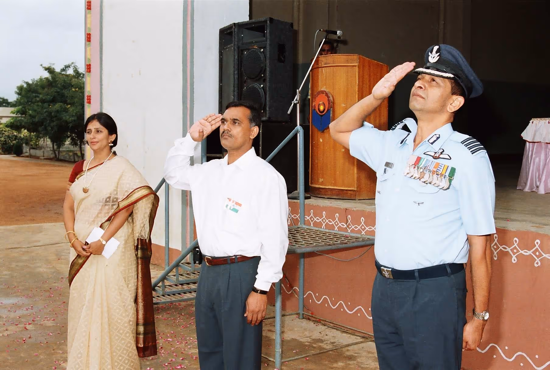 Two men saluting, one in an Air Force uniform with medals and the other in a white shirt with an Indian flag badge, standing next to a woman in a traditional sari.