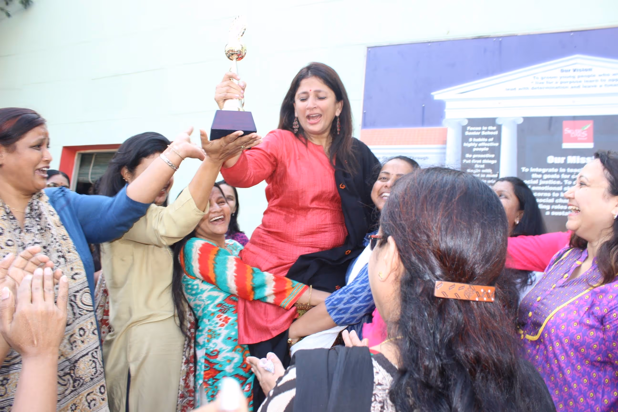 Group of women celebrating as one woman in a red top holds up a trophy while being lifted by others.