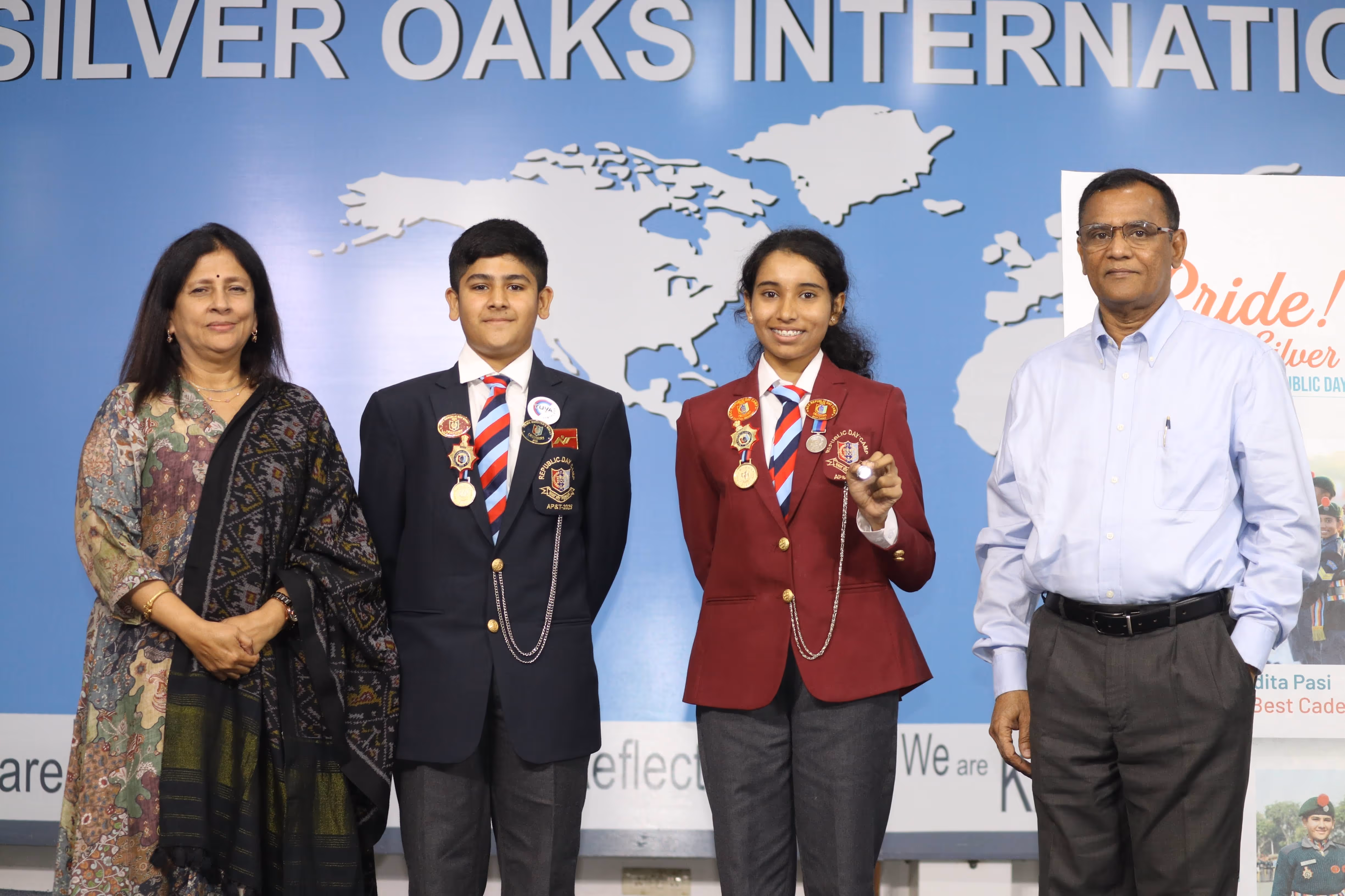 Two students in blazers with medals standing between a woman in a patterned shawl and a man in a light blue shirt in front of a blue wall with world map and Silver Oaks International text.